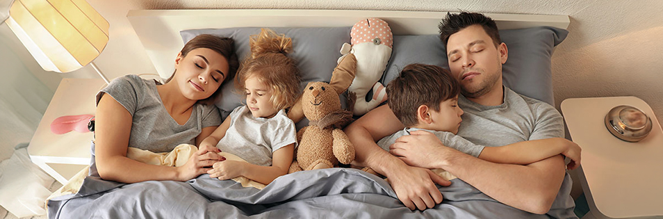 Vier Personen liegen gemeinsam in einem Bett, zwei Kinder halten Kuscheltiere in den Armen.