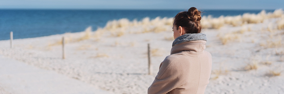 Person steht an einem Strand und blickt auf das Meer. Im Hintergrund sind Sanddünen zu sehen.