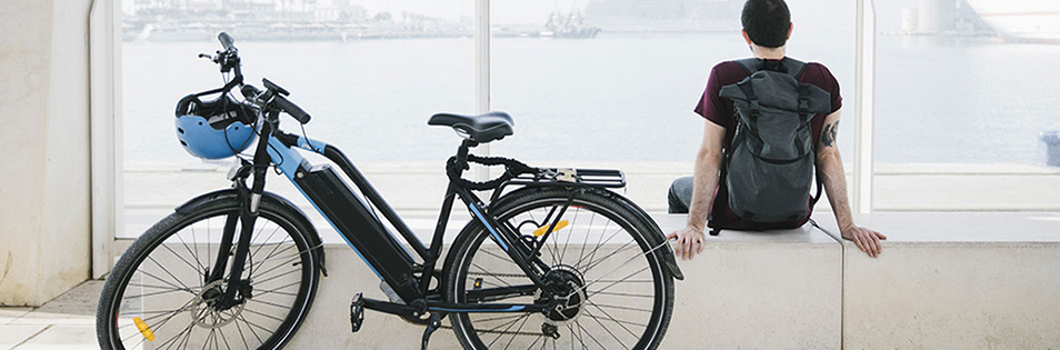 E-Bike mit Fahrradhelm steht neben einer sitzenden Person vor einer großen Fensterfront.