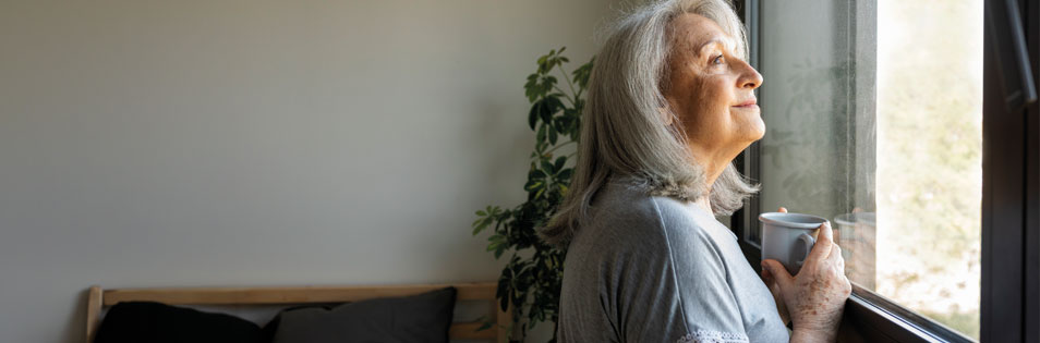 ältere Dame steht mit einer Kaffeetasse in der Hand vor dem Fenster und schaut zuversichtlich in die Ferne