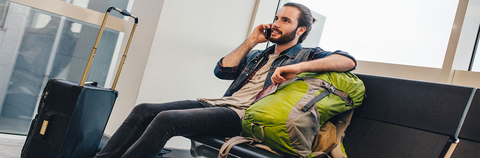 Person sitzt mit Rucksack und Rollkoffer in einem Wartebereich und telefoniert mit einem Mobiltelefon.