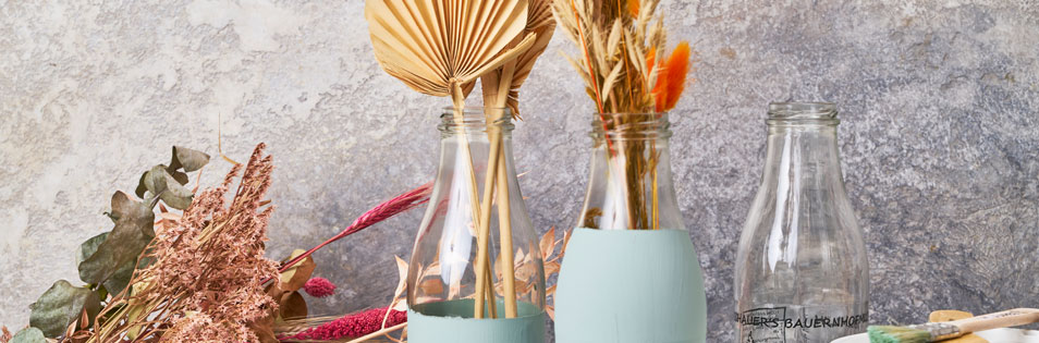 Glasflaschen mit Trockenblumen und dekorativen Pflanzen stehen vor einer strukturierten Wand. Die Flaschen werden als Vasen für die Dekoration genutzt