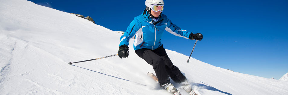 Person fährt mit Skiern auf einer verschneiten Piste unter blauem Himmel. Im Hintergrund sind schneebedeckte Berghänge zu sehen.