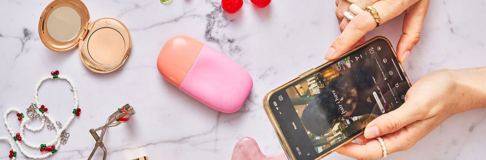 Auf einem hellen Marmortisch liegen Kosmetikprodukte, Schmuck und ein Smartphone, auf dessen Bildschirm eine Social-Media-Anwendung geöffnet ist.