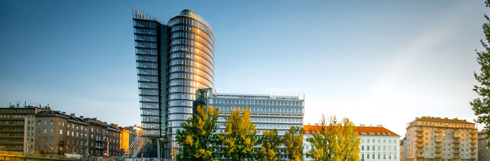 UNIQA Tower - Moderner Hochhausturm mit Glasfassade an einem Gewässer. Vor dem Gebäude stehen Bäume, im Umfeld befinden sich weitere Gebäude der Stadt.