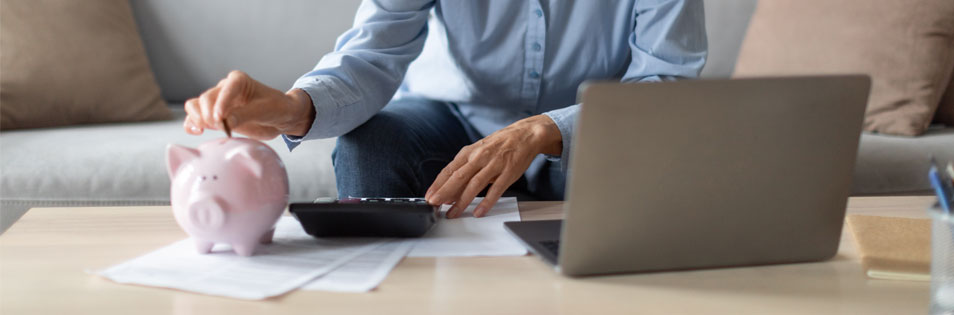 Person sitzt an einem Tisch und nutzt einen Taschenrechner. Auf dem Tisch liegen Unterlagen, daneben stehen eine Tasse und ein Laptop.