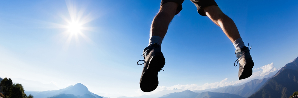 Person springt im Freien vor einer Berglandschaft und blauem Himmel.