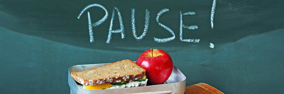 Jausenbox mit belegtem Brot und einem Apfel vor einer Tafel mit der Aufschrift „PAUSE!“