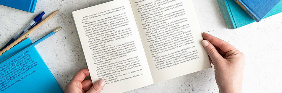 Hände halten ein aufgeschlagenes Buch auf einem Tisch mit weiteren Büchern und Schreibutensilien.