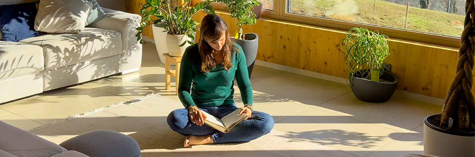 Person sitzt in einem hellen Wohnraum auf dem Boden und liest ein Buch