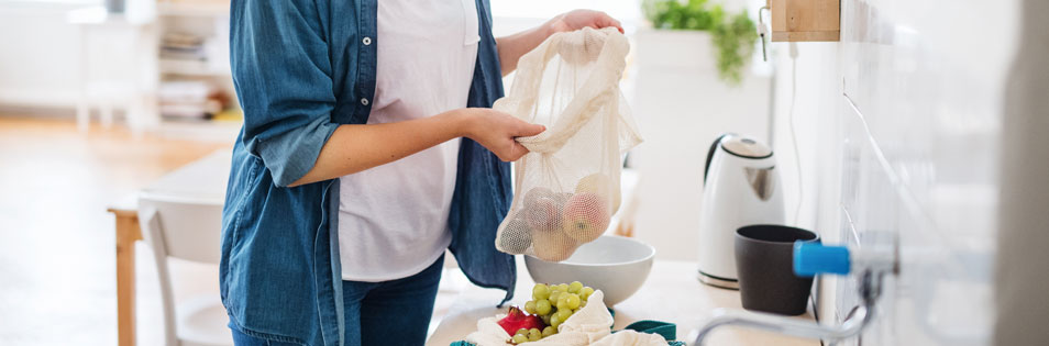 Person hält einen wiederverwendbaren Netzbeutel mit Obst über einer Küchenarbeitsfläche. Auf der Arbeitsfläche liegen weitere Lebensmittel und Stoffbeutel