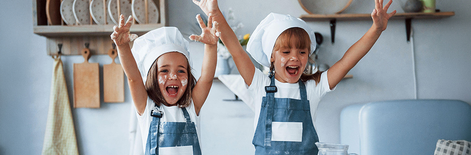 Zwei Kinder mit Kochmützen und Schürzen stehen in einer Küche mit erhobenen Armen vor einer Arbeitsfläche.