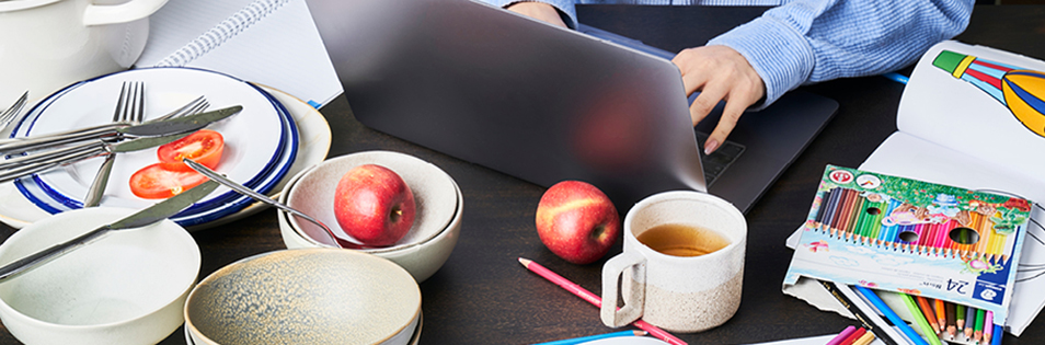 Arbeitsplatz mit Laptop, Geschirr, Obst und Malutensilien auf einem Tisch.
