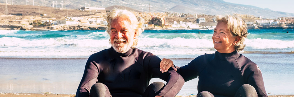Zwei ältere Personen sitzen in Sportbekleidung an einem Strand. Im Hintergrund sind Meer, Küste und Gebäude zu sehen.