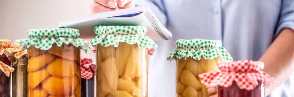 Mehrere Gläser mit eingekochtem Obst und Gemüse stehen auf einer Arbeitsfläche. Im Hintergrund macht sich eine Person Notizen in einem Notizblock.