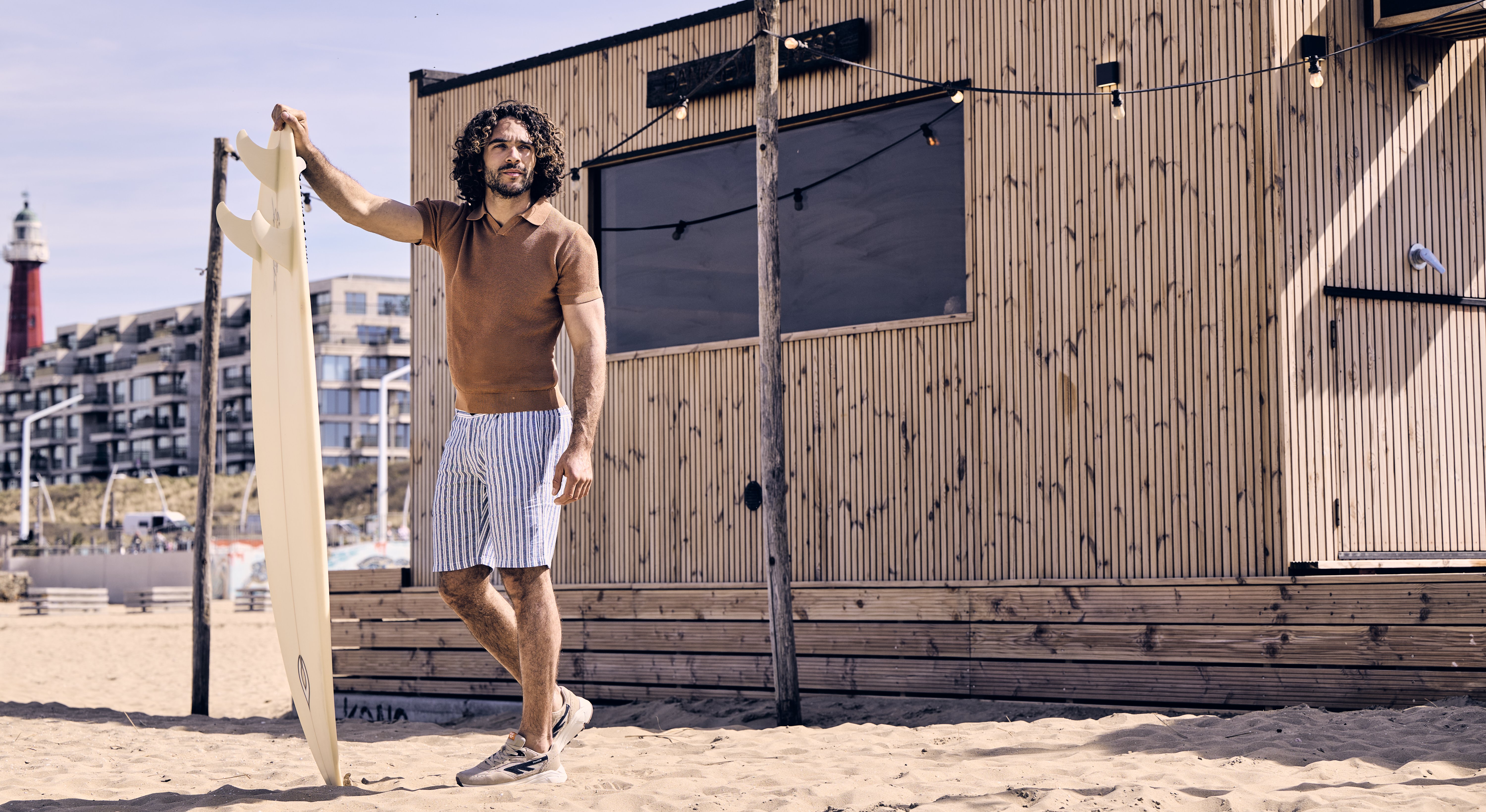 Man in casual beachwear holds a surfboard on sandy beach with a wooden building and lighthouse in the background.