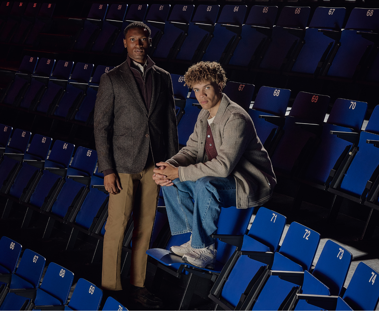 Two people in a theater with blue seats, one standing and one sitting, dressed in casual attire, looking towards the camera.