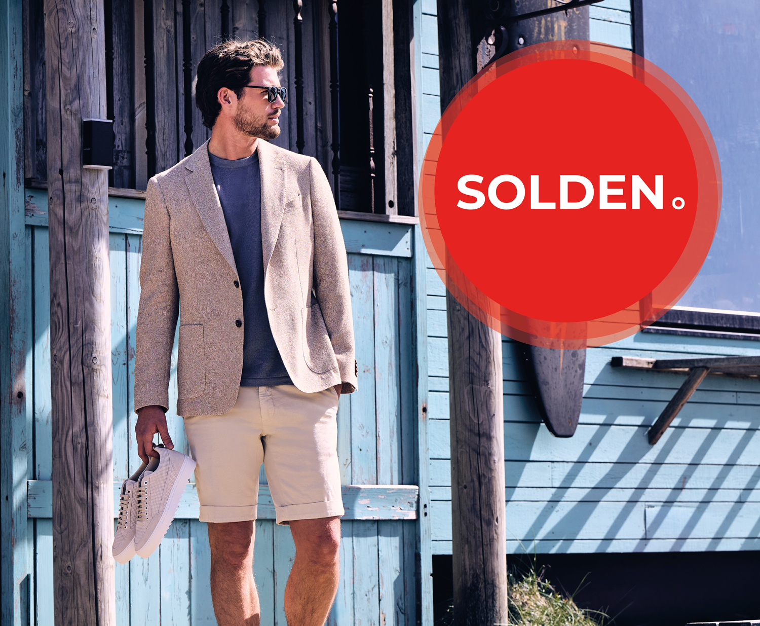Man in beige blazer and shorts holding hat, standing by a turquoise wooden structure. Large red circle with "SOLDEN" text overlaps the image.