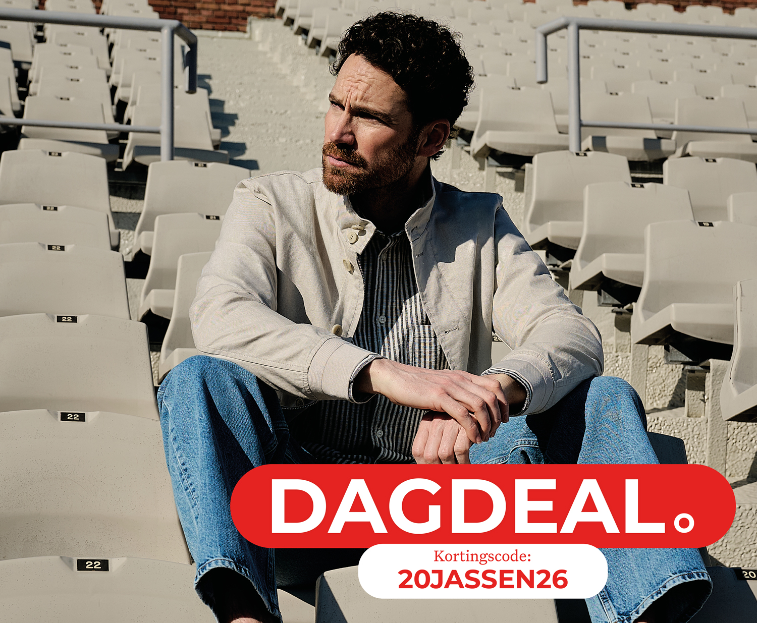 Man sitting on stadium seats wearing a light jacket and jeans. Text overlay: "DAGDEAL" with discount code "20JASSEN26."
