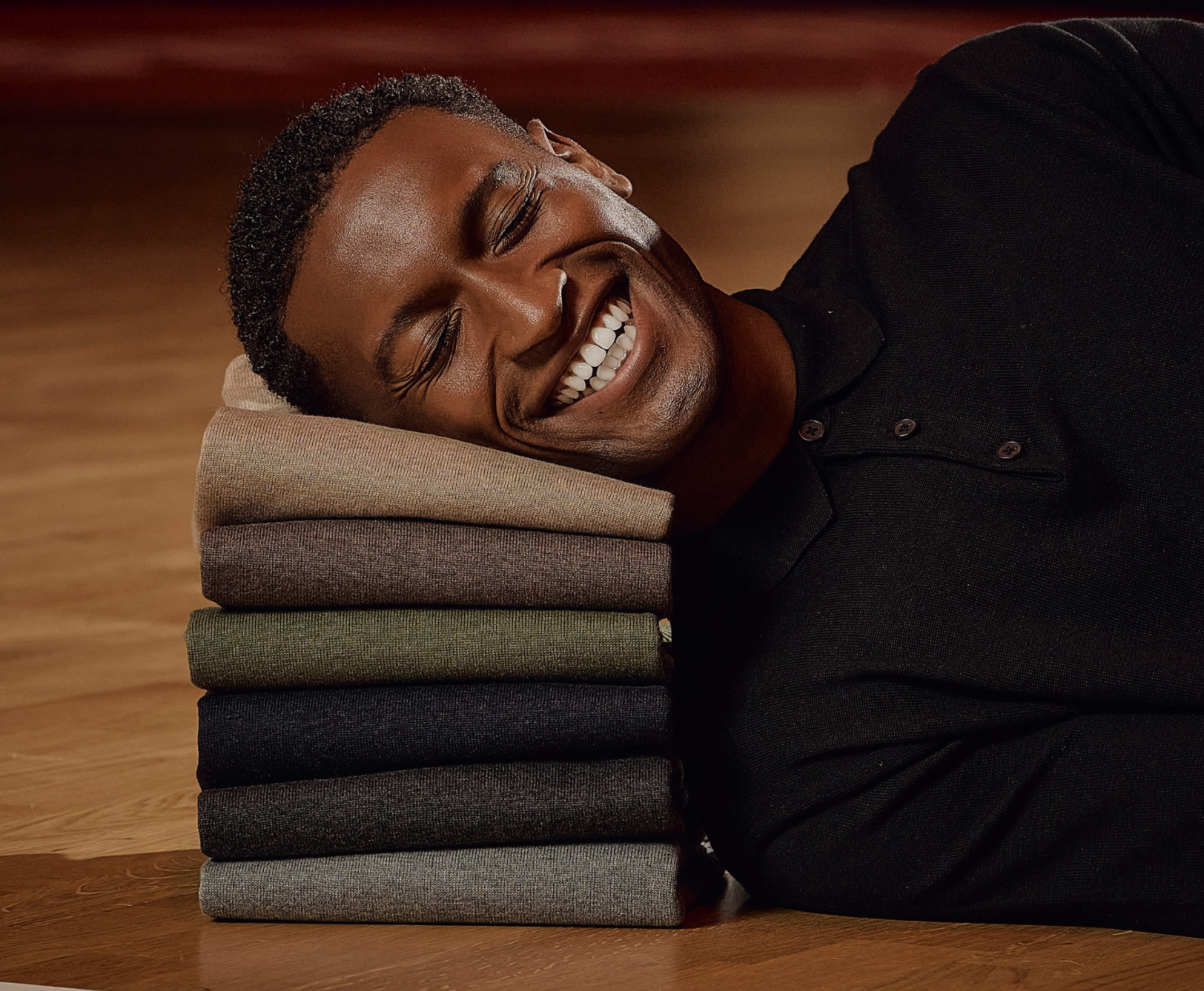A man smiles with his head resting on a colorful stack of folded sweaters, lying on a wooden floor.