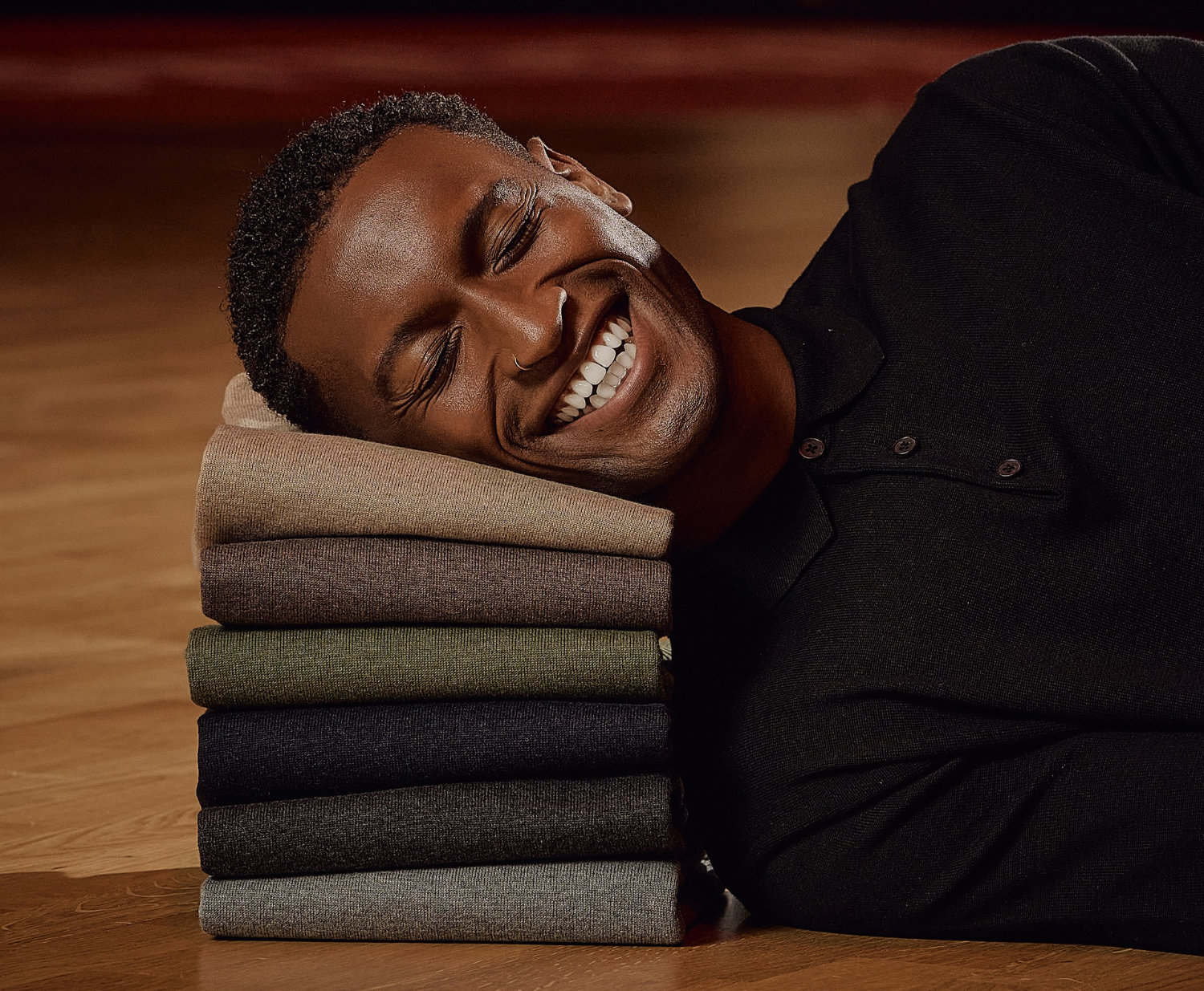 Man smiling with eyes closed, resting his head on a stack of colorful folded sweaters on a wooden floor.