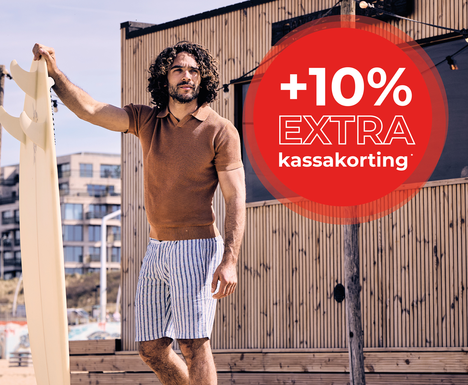 A man with curly hair stands by a surfboard, wearing a brown shirt and striped shorts, with a red "+10% EXTRA kassakorting" overlay.