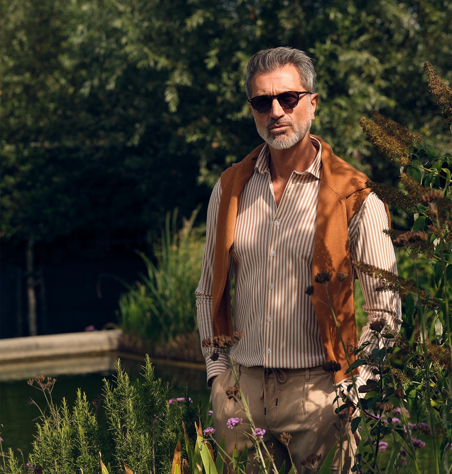 Man gekleed in een bruin overhemd met witte strepen en een schipperstrui geknoopt om zijn nek poserend in een tuin