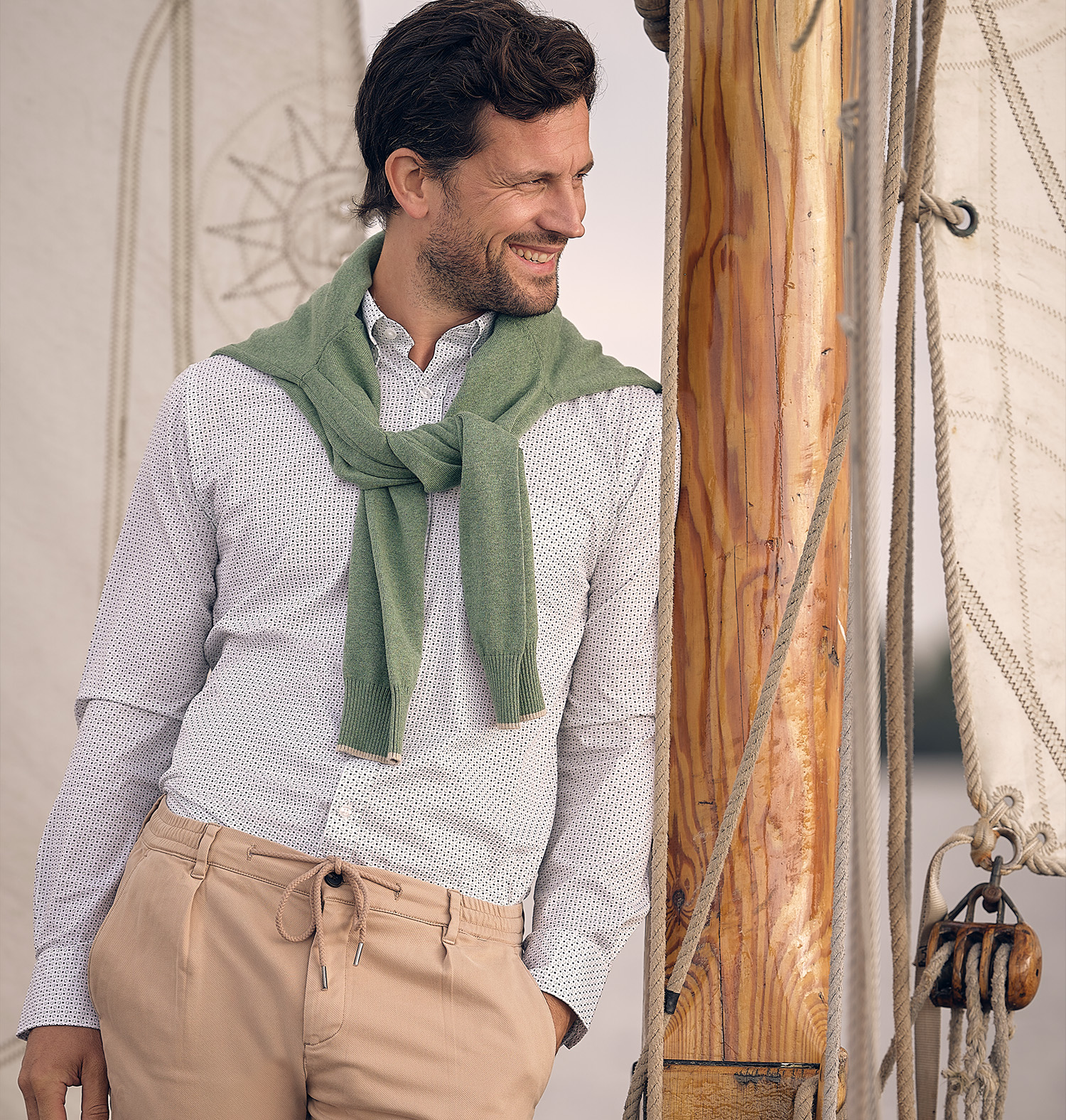 man gekleed in een overhemd, beige chino en heeft een groene schipperstrui omgeknoopt. Leunend tegen een mast op een boot