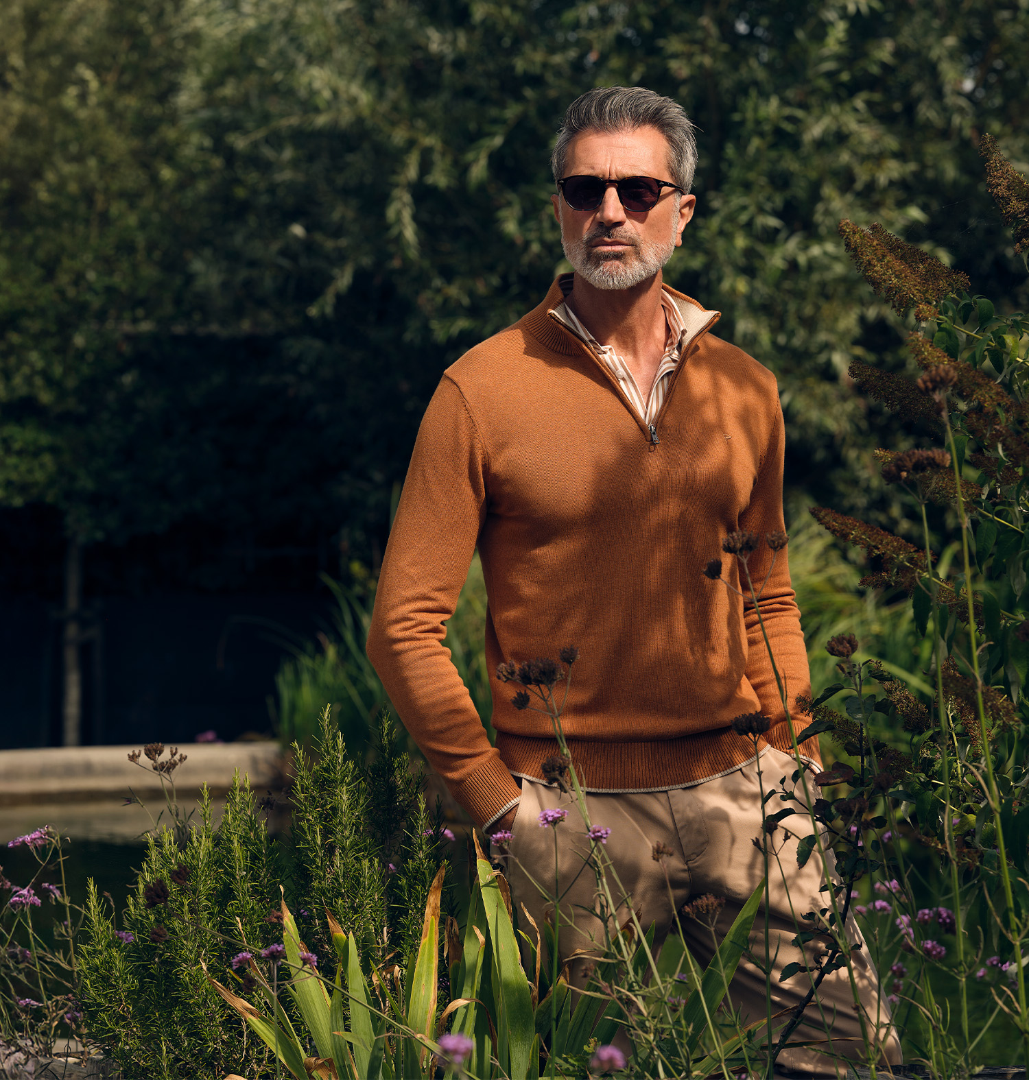 man poserend met een bruine trui en chino in een tuin met op de achtergrond een vijver