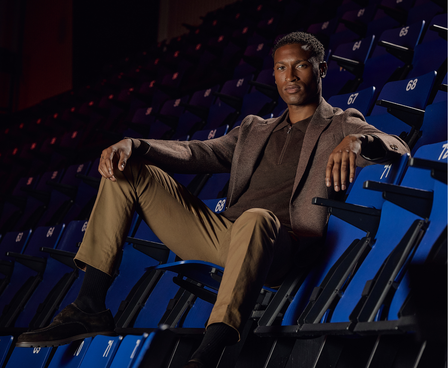A man in a brown blazer and khaki pants sits relaxed in blue stadium seats, with dark ambiance around.
