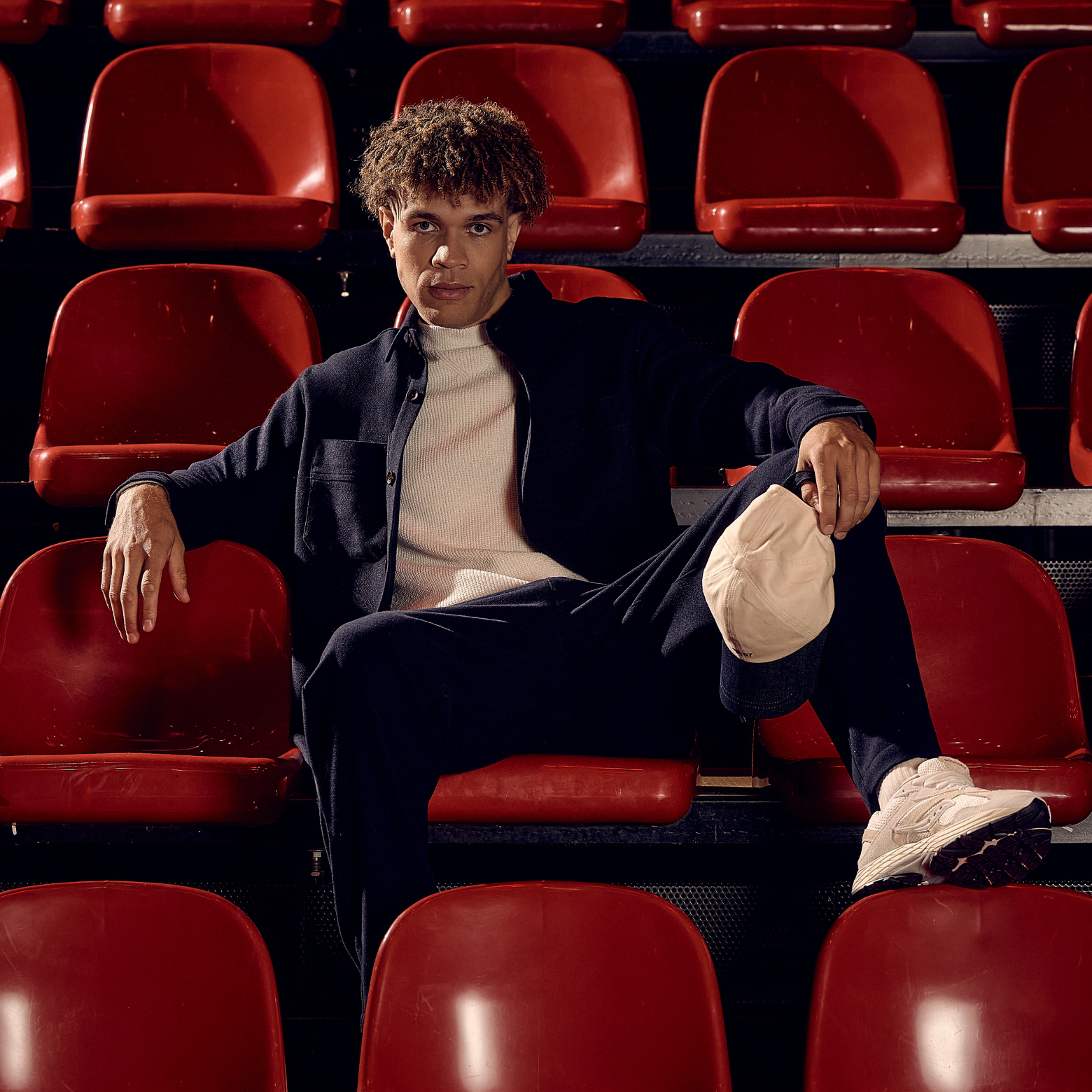A person in a dark outfit and sneakers sits casually on red stadium seats, with one leg crossed and arm resting on a chair back.