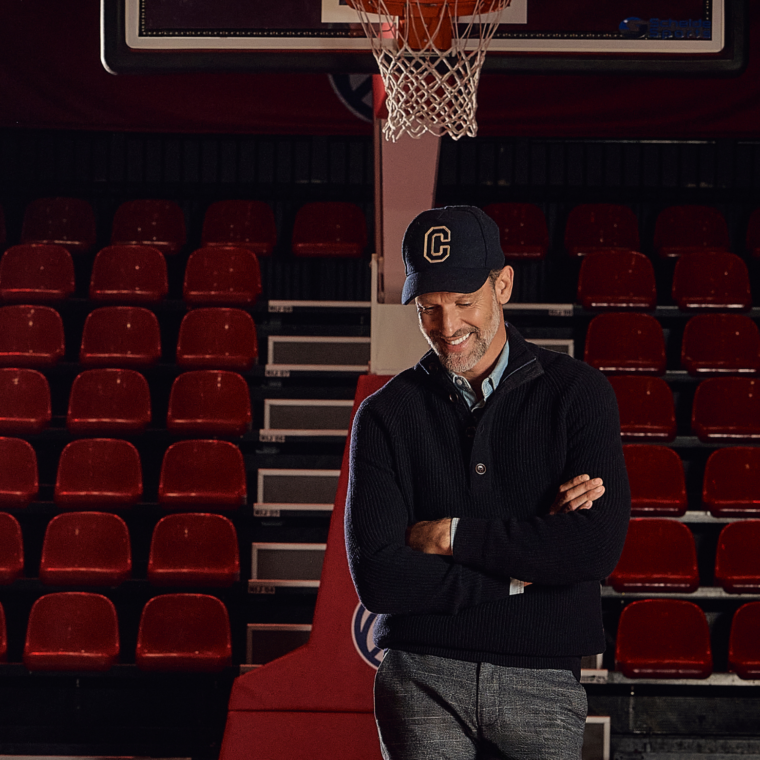 Man in a dark sweater and cap, standing with crossed arms on a basketball court, surrounded by empty red bleacher seats.