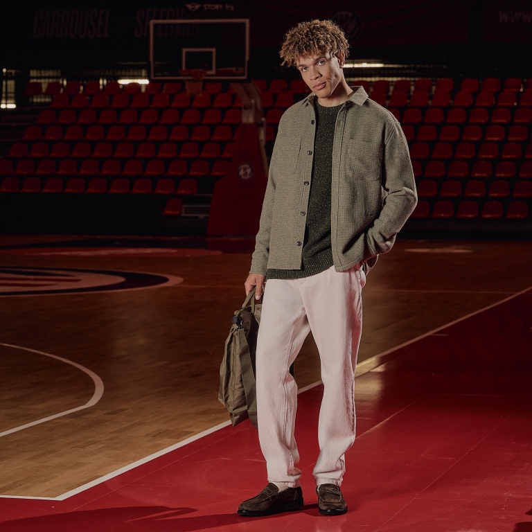 A person stands on a basketball court, wearing a green sweater, light jacket, white pants, and holding a bag, with red seats in the background.