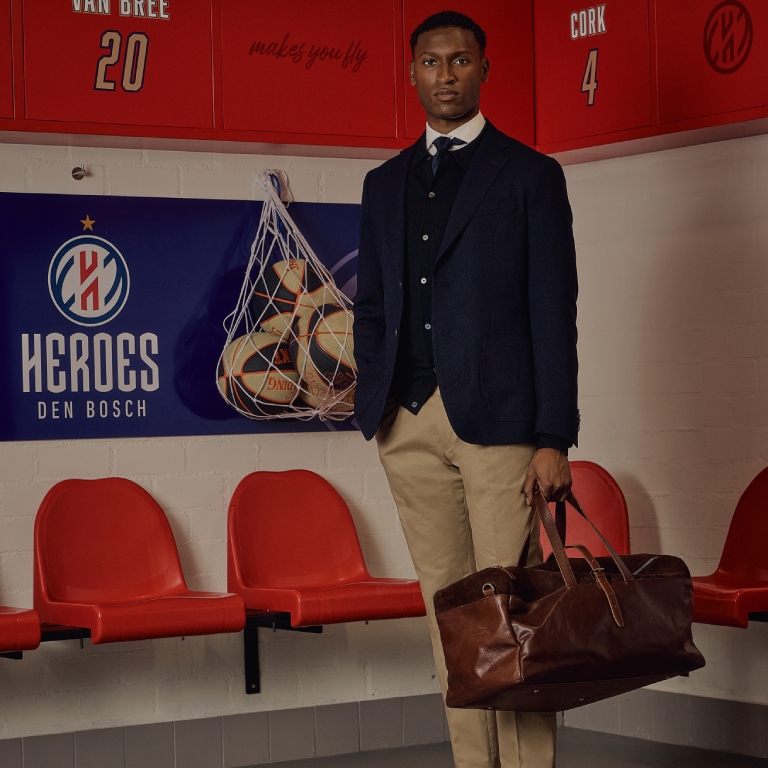 A man in a suit stands in a sports locker room holding a leather bag. Red seats and basketballs in netting are visible in the background.