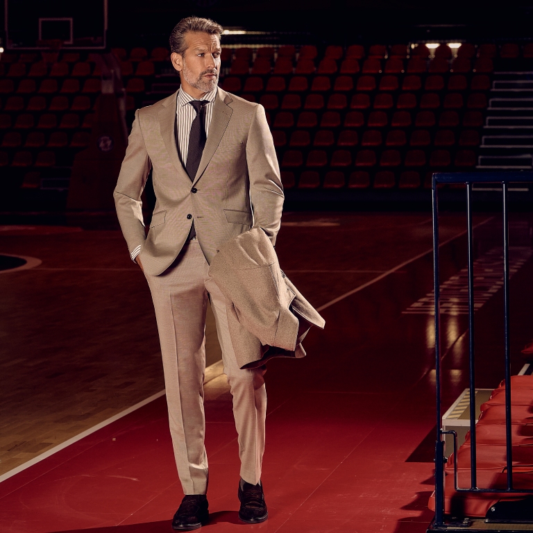 A man with gray hair wearing a beige suit and holding a coat walks in an empty basketball arena with dim lighting.