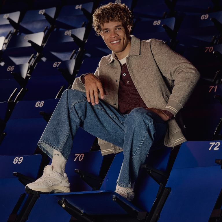 Man with curly hair sits casually on stadium seats, wearing a beige jacket, maroon shirt, blue jeans, and white sneakers.