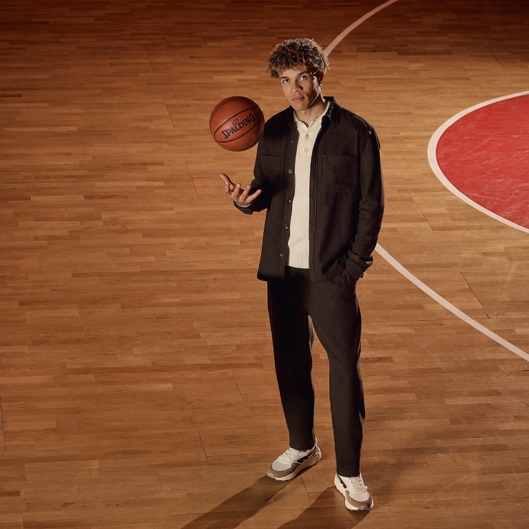 A person stands on a basketball court, spinning a basketball on their finger, wearing casual attire with white sneakers.