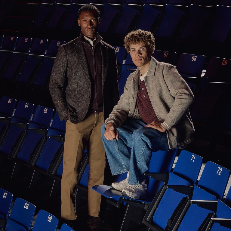 Two people, one standing and one sitting on the armrest of a blue stadium seat, both wearing jackets, looking at the camera.