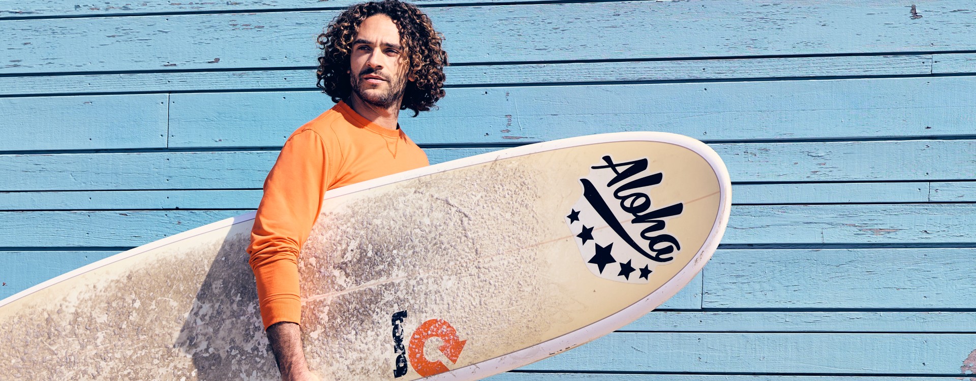 Man in orange shirt holding surfboard with "Aloha" text, standing against a blue wooden wall.