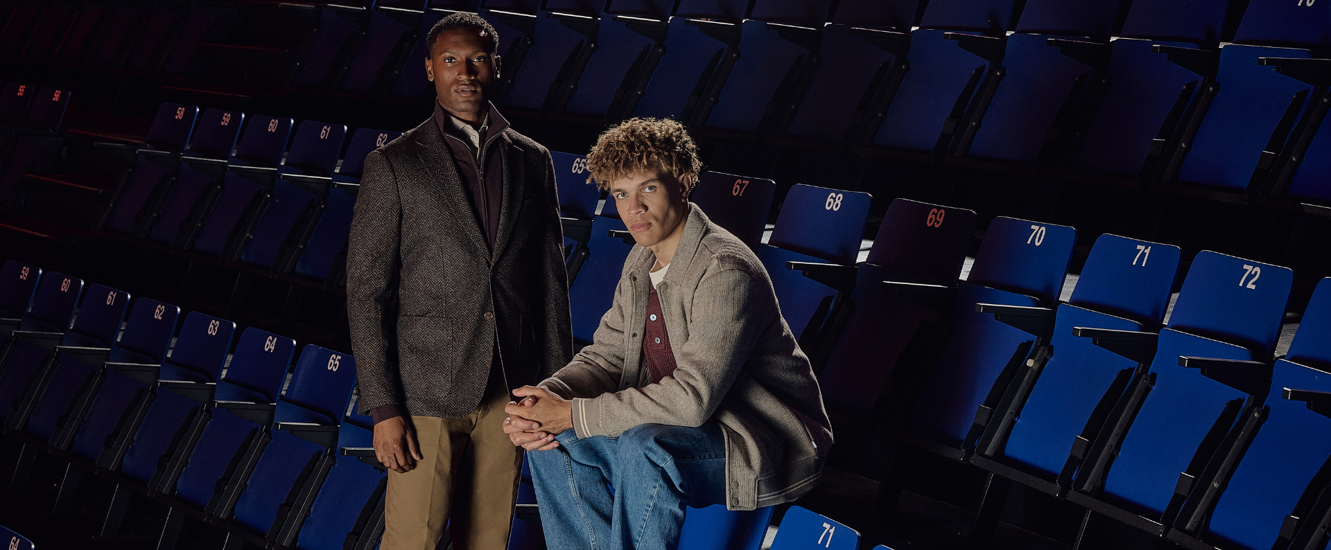 Two men in a theater with blue seats; one stands wearing a dark suit, the other sits casually in a light jacket and jeans.
