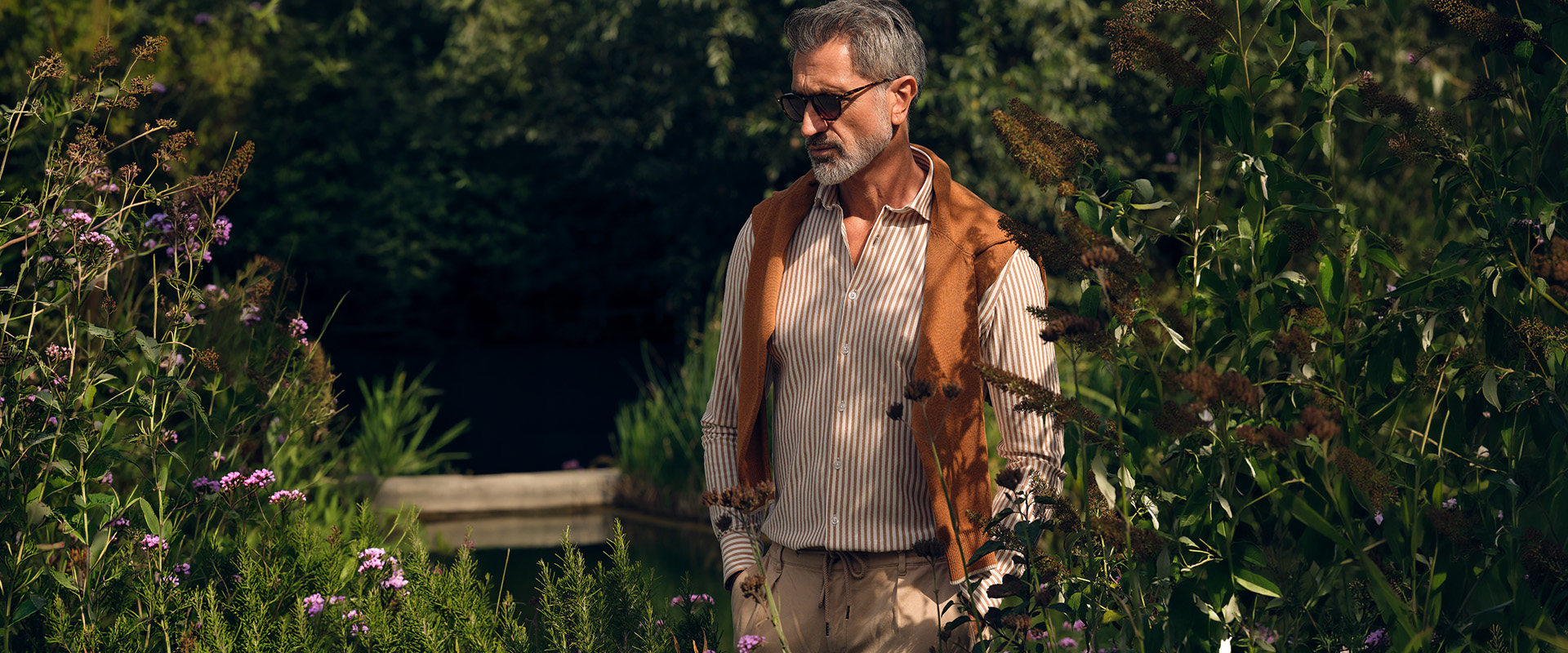 Man gekleed in een bruin overhemd met witte strepen en een schipperstrui geknoopt om zijn nek poserend in een tuin