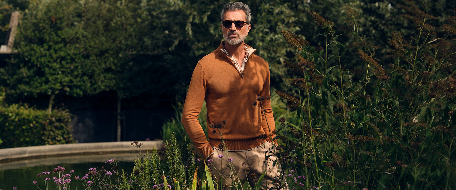 man poserend met een bruine trui en chino in een tuin met op de achtergrond een vijver