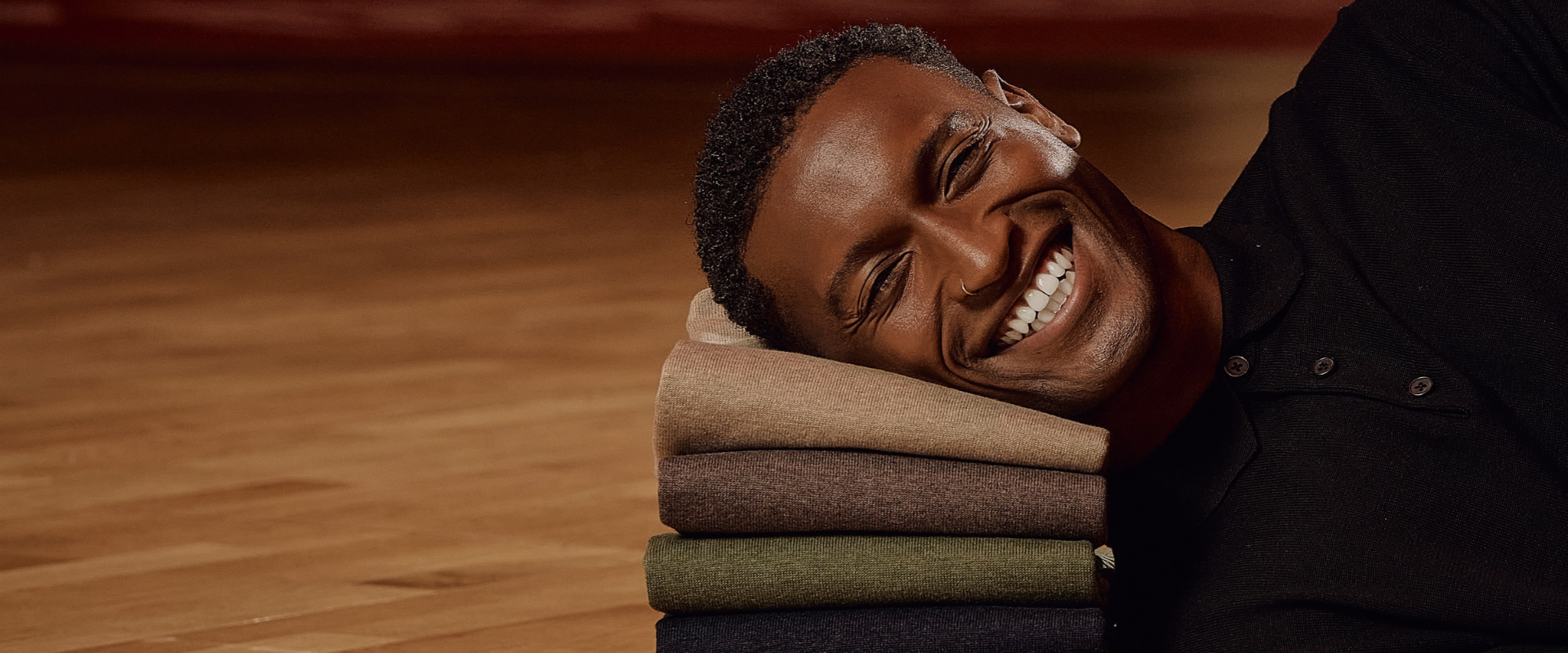 Smiling man lying on a wooden floor, resting his head on a stack of folded sweaters in tan, brown, green, and black.