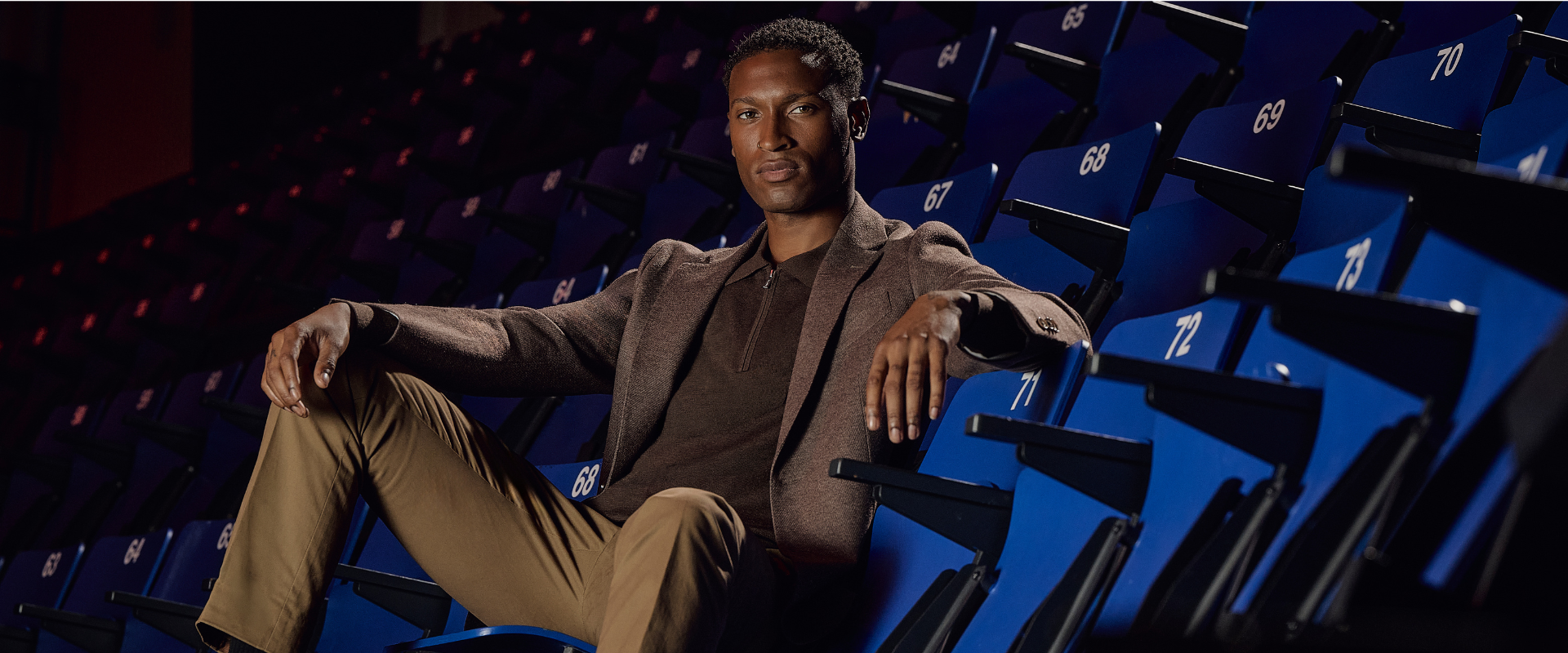 A person sits relaxed in a row of blue stadium seats, wearing a brown blazer and tan pants, with numbers marked on the seats around them.