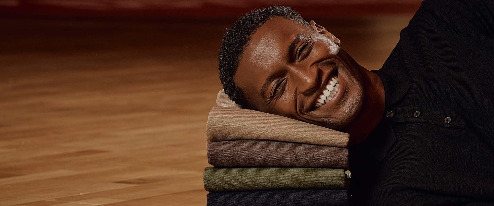 A smiling man lies on a wooden floor, resting his head on a stack of folded clothes in earthy tones.