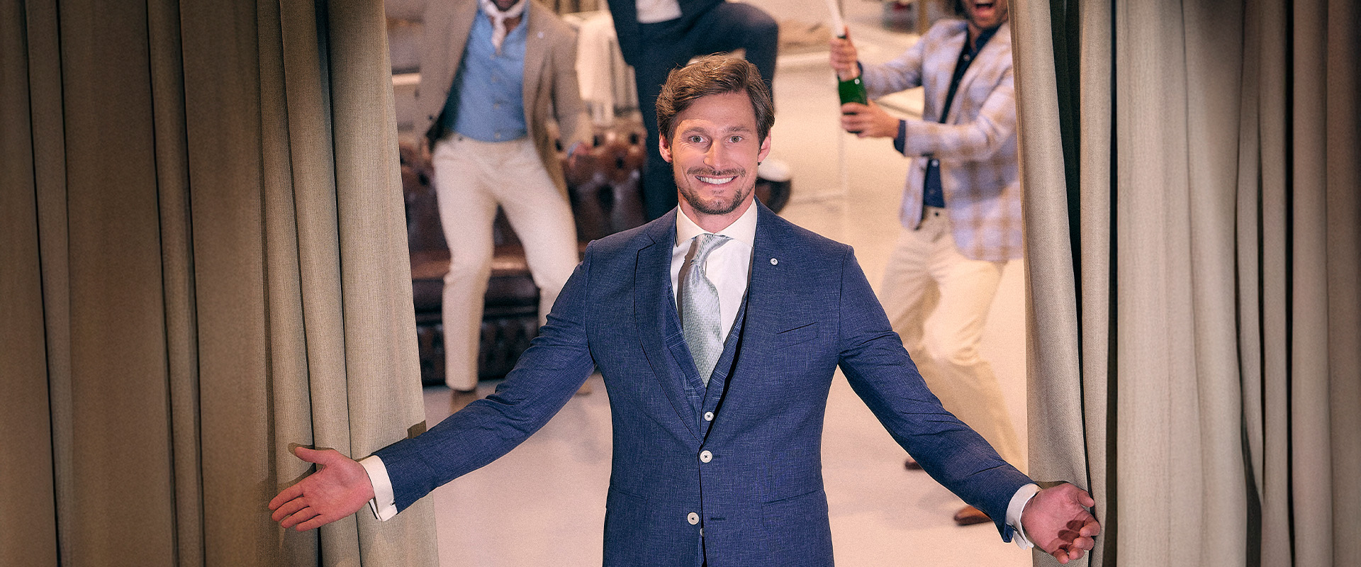 Model met open armen in een blauw trouwpak, op de achtergrond blije mannen