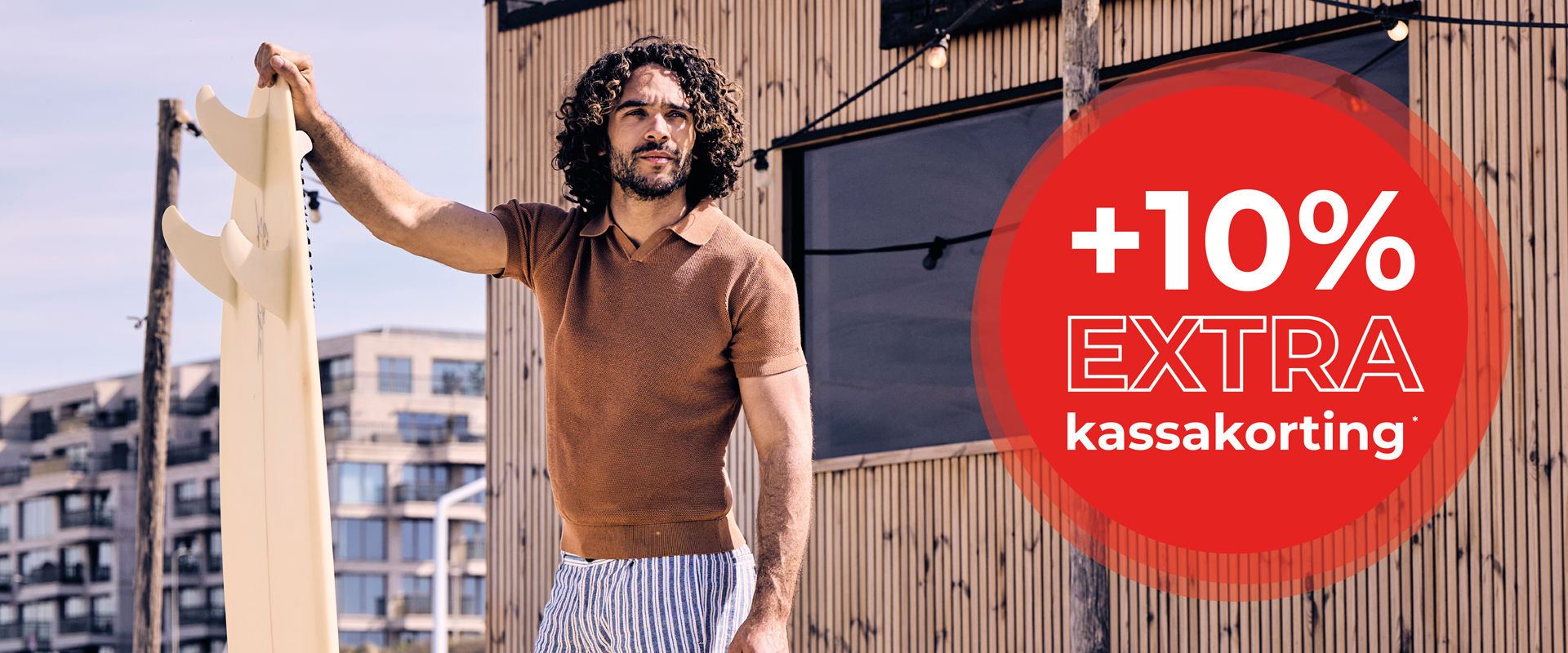 Man in a brown shirt holding a surfboard, standing outside with a building in the background. A red circle shows "+10% EXTRA kassakorting."