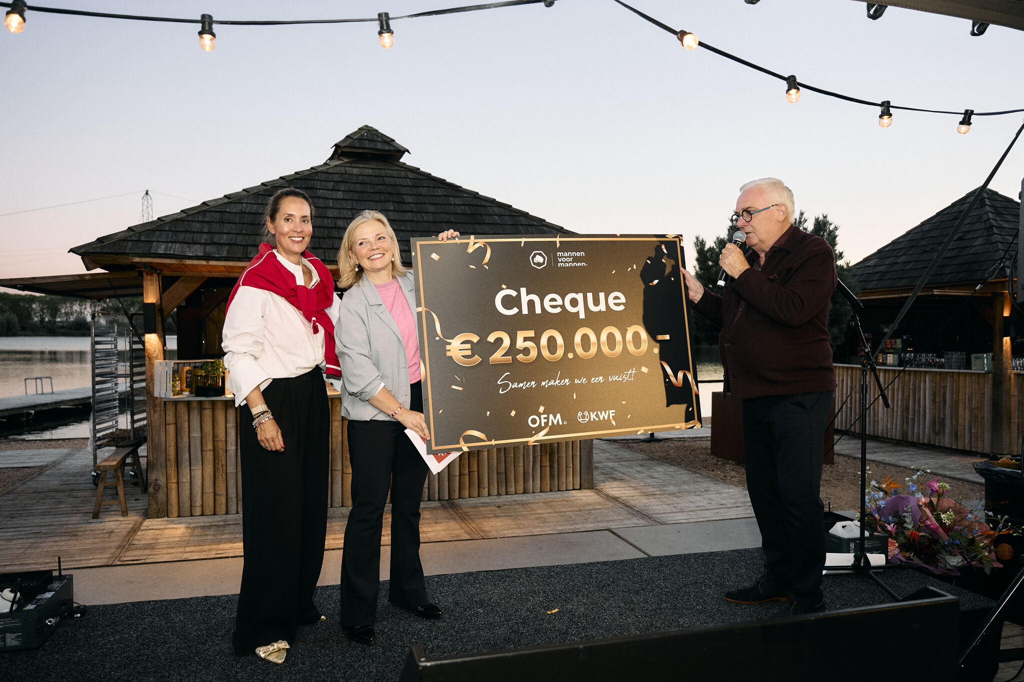 Three people on stage hold a large cheque for €250,000, with festive lighting and a wooden gazebo in the background.