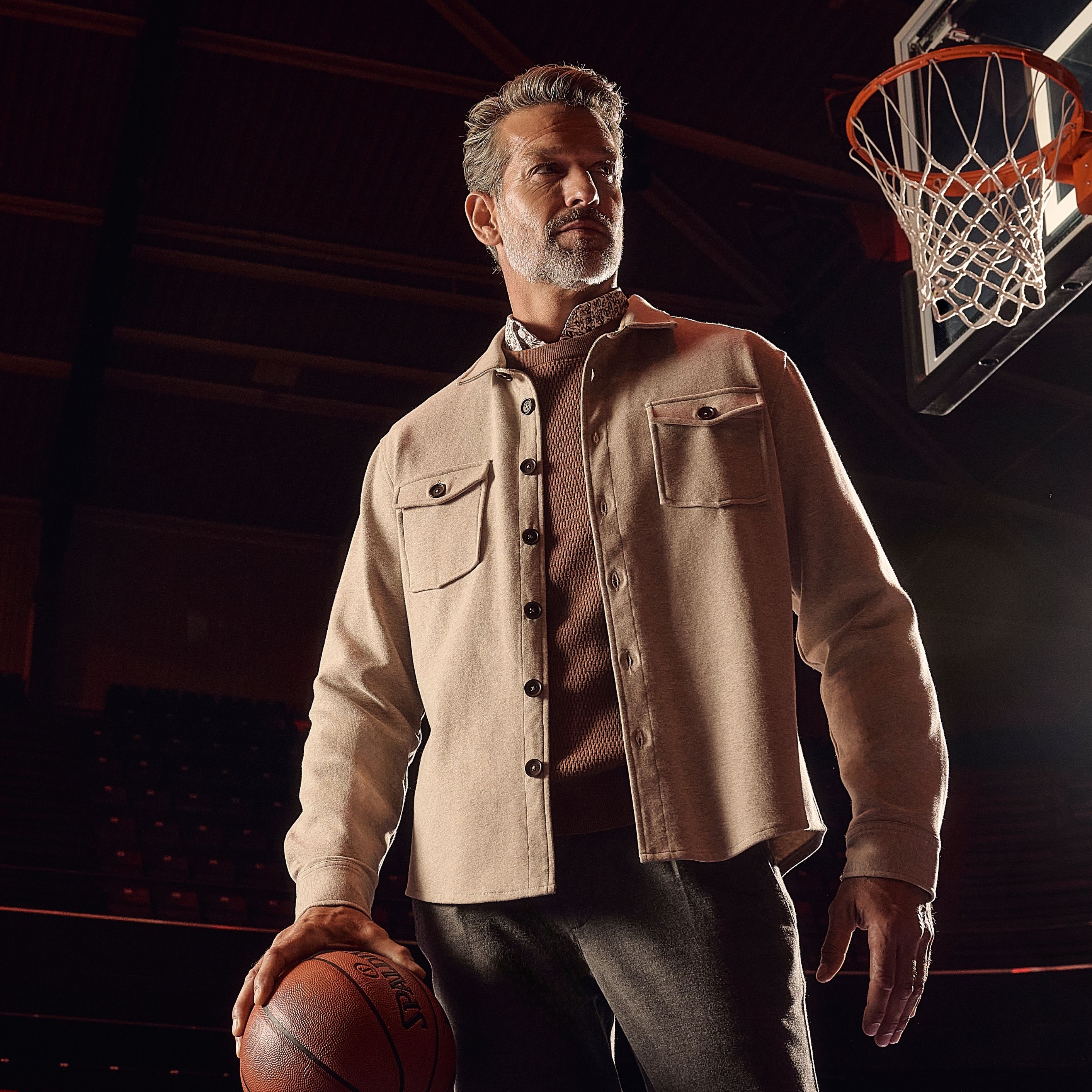 Man stands on a basketball court with a ball, under a hoop, wearing a beige jacket and dark pants.