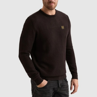 Man wearing a dark long-sleeve sweater with a small emblem on the chest, paired with dark jeans, against a light gray background.